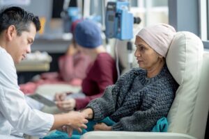 A healthcare professional checks on a patient wearing a headscarf while the patient sits in an infusion chair receiving treatment at a medical clinic. Another patient receiving treatment is visible in the background.
