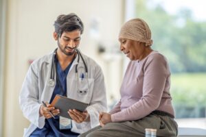 A healthcare professional reviews information on a tablet with a patient wearing a headscarf seated on an exam table during a medical appointment.