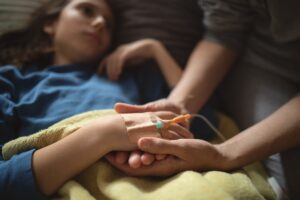 Close-up of a child resting under a blanket with an intravenous (IV) line secured to the hand. Another person gently holds the child's hand, highlighting supportive care during medical treatment.