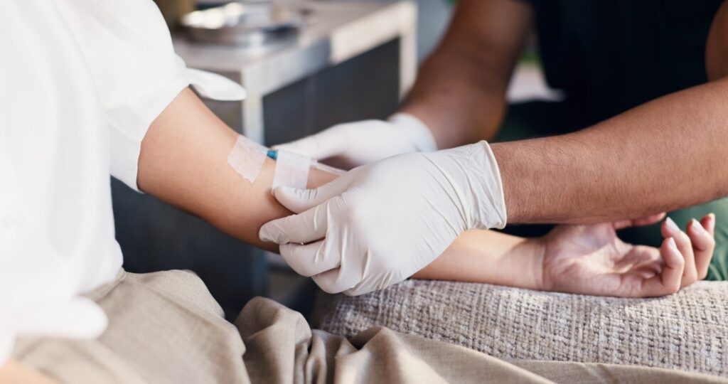 Close-up of a healthcare professional wearing protective gloves securing an intravenous (IV) cannula in a patient’s arm. The patient is seated, and medical tape holds the IV line in place during treatment or care.