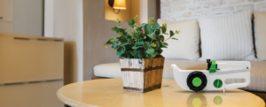 Freedom60 infusion pump resting on a table in a home setting beside a potted plant. The portable infusion device is displayed in the foreground, illustrating home-based infusion therapy in a comfortable domestic environment.