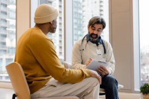 Healthcare professional with a stethoscope reviews notes on a clipboard while seated across from a patient in a consultation room with large windows and an urban skyline visible in the background.