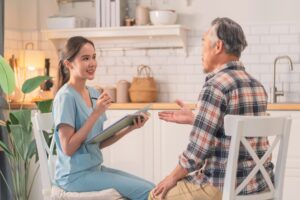 Healthcare worker in medical scrubs sits with an older adult at home, taking notes on a clipboard during a care assessment or wellbeing visit. The conversation takes place in a bright kitchen setting with household furnishings visible in the background.