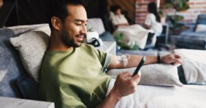 Person seated in a comfortable treatment chair looks at a smartphone while receiving intravenous therapy through an IV line in the arm. The setting appears to be an outpatient treatment or infusion area, with other people and seating visible in the background.