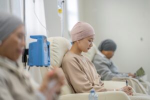 Person wearing a light pink head covering sits in a recliner receiving intravenous treatment in a clinical setting. Medical infusion equipment is visible beside the chair, with other patients seated in the background.