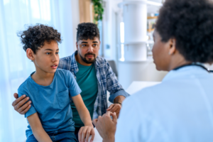 A child sits beside an accompanying adult while speaking with a healthcare professional during a consultation in a bright medical clinic. The healthcare professional is seated opposite the child, and the adult sits nearby with a hand resting on the child’s shoulder.