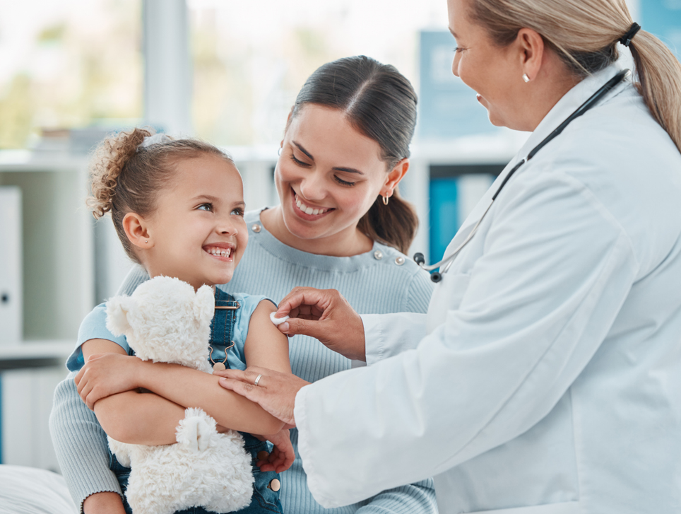 A healthcare professional in a white coat places a bandage on a young child’s upper arm while the child sits on an adult’s lap holding a white teddy bear in a bright medical clinic.