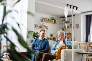 Healthcare professional in blue scrubs sitting with an older patient in a bright home care setting. An IV drip stand is positioned beside the sofa, with houseplants, shelves, and home furnishings visible in the background, illustrating in-home medical support and patient care.