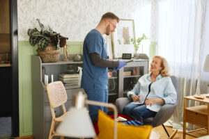 Healthcare worker in blue scrubs providing in-home care to an older adult seated in a chair. An IV drip stand and medical equipment are visible in the room, alongside bookshelves, houseplants, and modern furnishings, highlighting personalised healthcare support in a home environment.