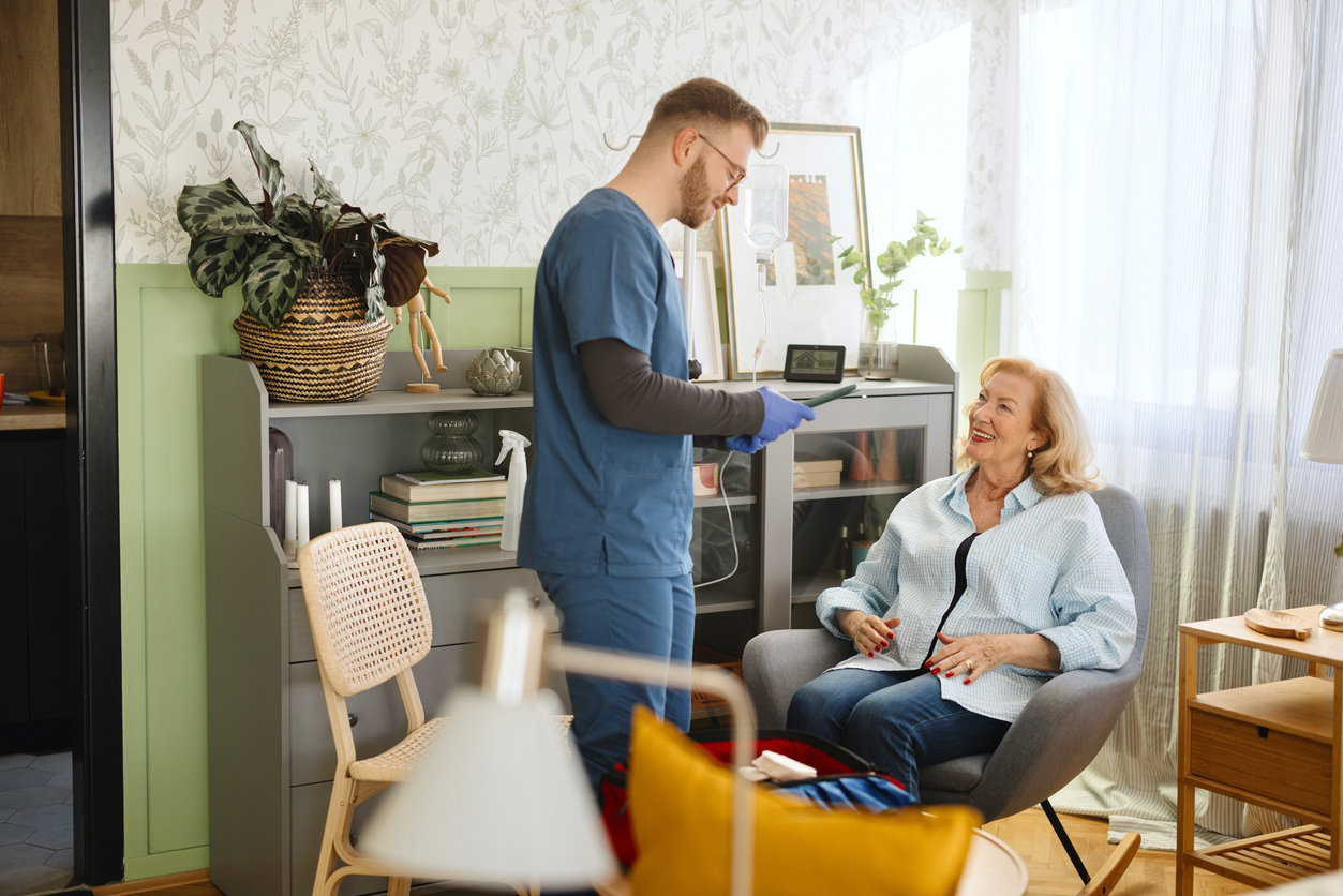 Healthcare worker in blue scrubs providing in-home care to an older adult seated in a chair. An IV drip stand and medical equipment are visible in the room, alongside bookshelves, houseplants, and modern furnishings, highlighting personalised healthcare support in a home environment.