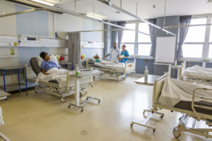 Hospital ward with multiple patient beds, bedside tables, and medical equipment. Two healthcare professionals stand beside a patient bed near large windows, while another patient rests in a bed in the foreground. The room is clean, bright, and spacious with privacy curtains and overhead support rails.