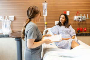 Patient seated in a hospital bed wearing a hospital gown while a healthcare professional holds a clipboard and speaks with the patient. The modern hospital room includes an IV drip, medical gas outlets, bedside equipment, and wood-panelled walls.